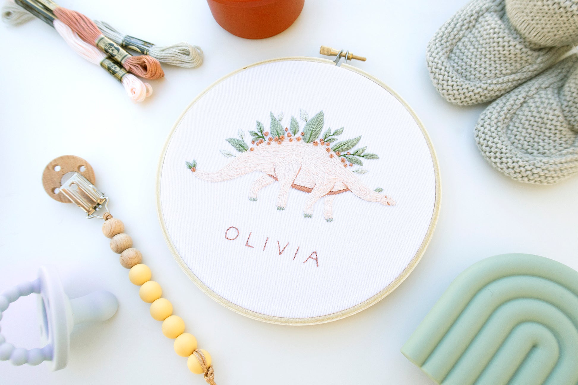 Pink dinosaur embroidery with leaves and flowers and custom baby name embroidery pattern on boho baby flat lay