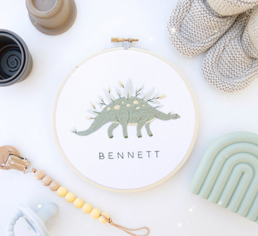 Green dinosaur embroidery with leaves and flowers and custom baby name embroidery pattern on boho baby flat lay