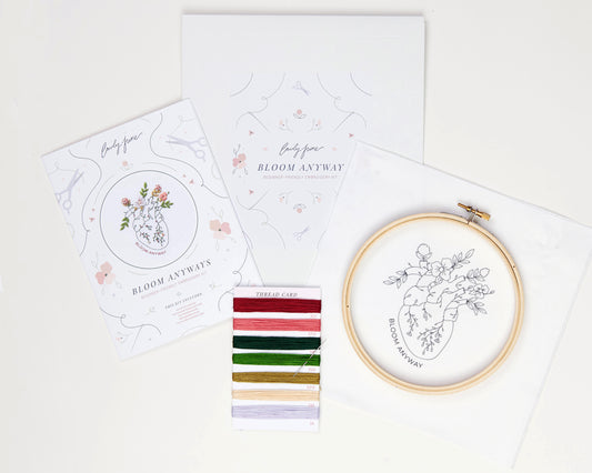 Bloom floral heart anatomy hand embroidery kit for mental health with pink, purple and red flowers and green leaves with DMC embroidery floss, printed embroidery fabric, embroidery needle, embroidery hoop and embroidery guide laid out on background