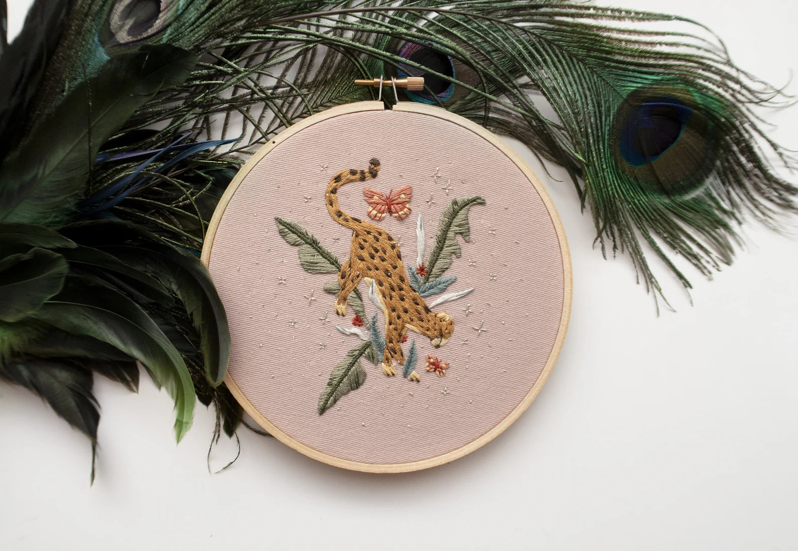 Cheetah in jungle hand embroidery pattern on light pink fabric, green jungle leaves, pink butterflies and silver stars with peacock feather