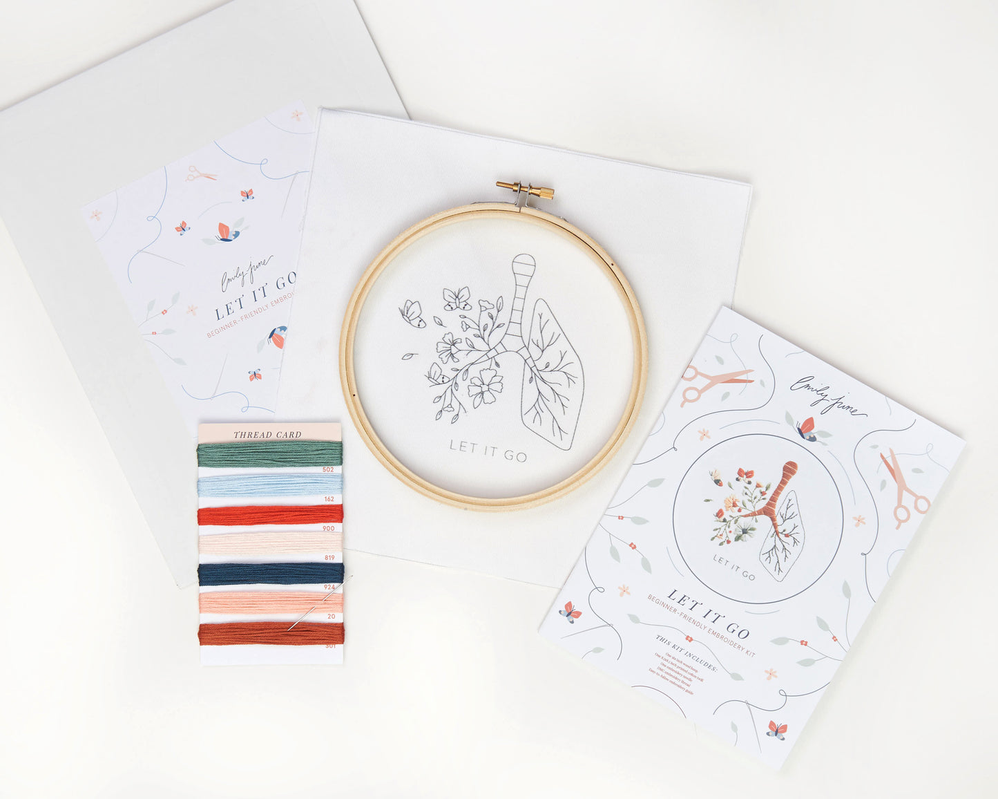 Let it go floral lung hand embroidery kit with pink and blue flowers and butterflies for mental health craft flat lay with printed embroidery fabric, wood embroidery hoop, embroidery guide, embroidery floss, and embroidery needle, kit contents laid out on white background