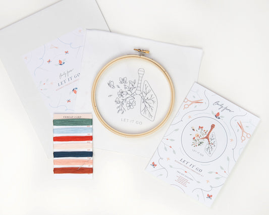 Let it go floral lung hand embroidery kit with pink and blue flowers and butterflies for mental health craft flat lay with printed embroidery fabric, wood embroidery hoop, embroidery guide, embroidery floss, and embroidery needle.