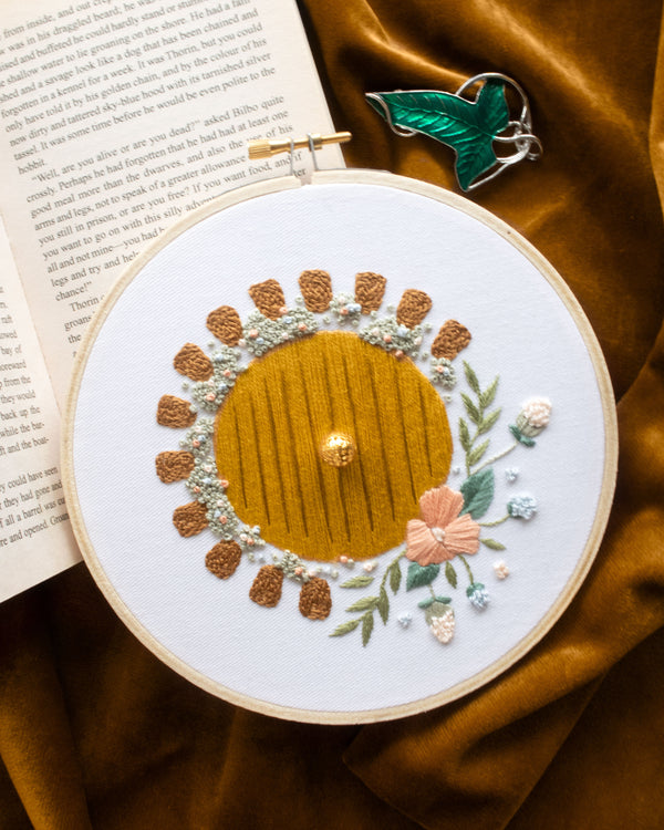 Hobbit Hole Embroidery Kit finished on dark yellow velvet background and green leaf cloak clasp and open book