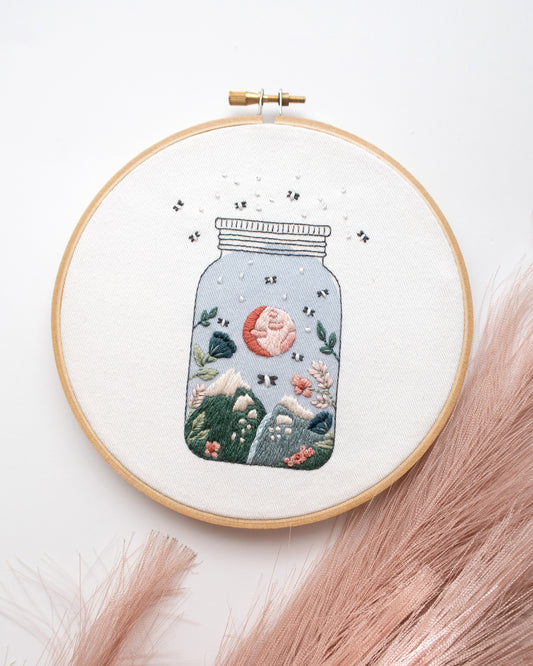 Fireflies, moon, mountain and flowers in magical jar hand embroidery kit on embroidery hoop with pink, blue, and green embroidery floss