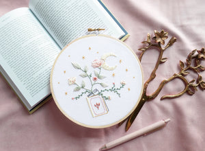 Embroidery pattern with open floral book with garden flowers and branches growing from open pages with stars and moon hand embroidery kit on embroidery hoop with boho dried florals on open book with floral scissors