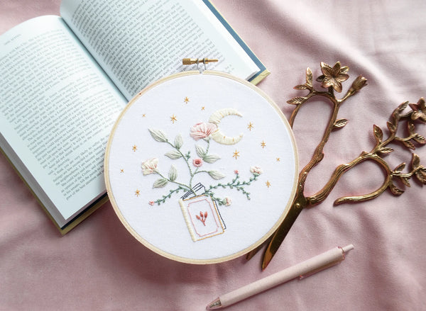 Embroidery pattern with open floral book with garden flowers and branches growing from open pages with stars and moon hand embroidery kit on embroidery hoop with boho dried florals on open book with floral scissors