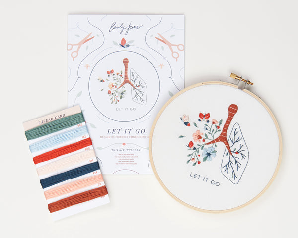Let it go floral lung hand embroidery kit with pink and blue flowers and butterflies for mental health craft flat lay with printed embroidery fabric, wood embroidery hoop, embroidery guide, embroidery floss, and embroidery needle.